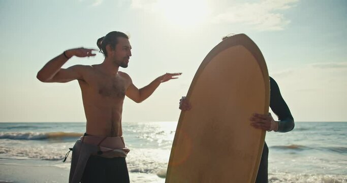 A brunette guy with a naked torso talks about his impressions of swimming on the Surf to his friend, a surfer in a wetsuit with a yellow longing for surfing on the seashore in the summer