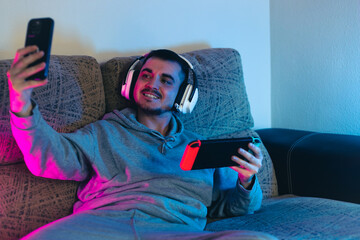 young blogger playing the console on his gaming set up illuminated with pink and blue light, talking on voice chat with white headphones with his cyber friends