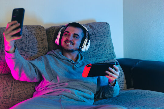 young blogger playing the console on his gaming set up illuminated with pink and blue light, talking on voice chat with white headphones with his cyber friends