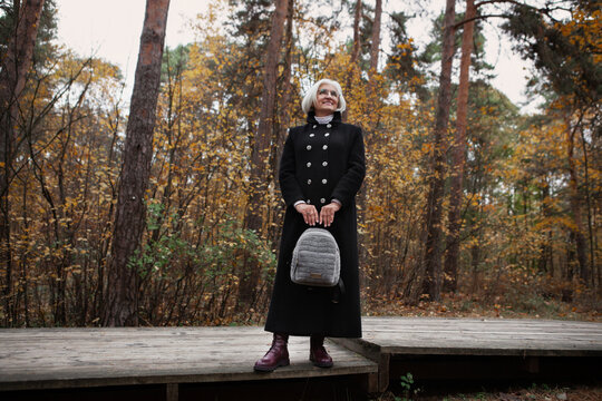 Refined older woman with silver hair stands in autumn forest, enjoys tranquil beauty of nature. Beautiful elderly female model in black coat. Mature joy, natural relaxation, and timeless beauty