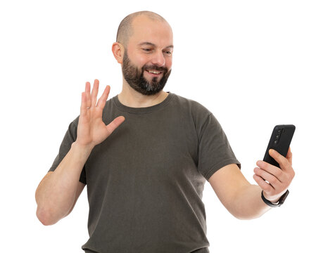 Portrait oh happy man with beard wearing brown t-shirt take selfie and waving raised palm, isolated on white background. Bald guy holding cell phone and say HI, HELLO, greeting you, welcoming guests.