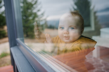 Sweet toddler child put his hands on the window. A little happy boy is looking outside through the...