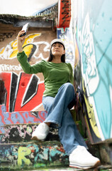Fototapeta premium Asian female tourist in casual clothing style using mobile phone taking selfie photo while sitting on stairs on open graffiti sprayed wall art,Street Fashion Woman wearing Green Long Sleeve,Blue Jeans