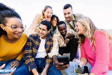 Young group of happy people having fun using mobile phone at city street. Smiling multiracial friends sitting together outside watching social media content on cellphone app. Gen tech people concept