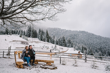 Couple in love with blanket hugs in winter on hill near wood forest. Man and woman holding cup drinking coffee. Recreation area, fire pit, fireplace in backyard house. Cabin terrace with mountain view