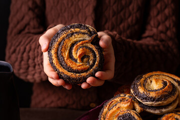 Girls hands holding homemade poppy seeds bun, swirl shape. Close up. Dark brown background.