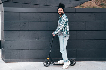 Trendy smiling bearded man in casual clothes riding electric scooter in urban background. Handsome model posing in the street near wall. Hipster guy with curly hairstyle. Cheerful and happy