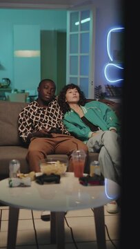 Vertical pan shot of diverse bored couple sitting on couch and watching TV using remote in neon lit room