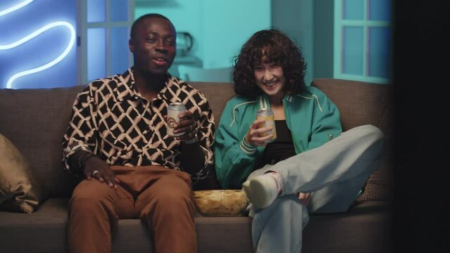 Medium shot of diverse couple laughing and drinking soda while spending time together watching TV on couch in neon lit living room