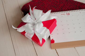Red gift box and calendar on  isolated on a red knitted scarf with white wooden background with. Christmas's day concept. Planning scheduling agenda, Event, organiser x'mas day. Flat lay, top view.