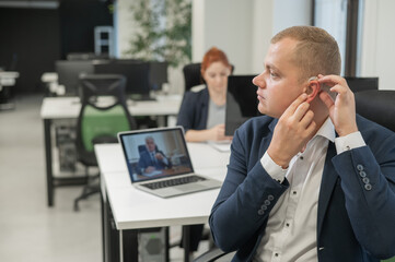 Caucasian man putting on hearing aid to online meeting on laptop. 