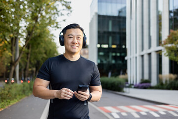 Smiling young Asian man standing on city street, running wearing headphones and holding phone,...