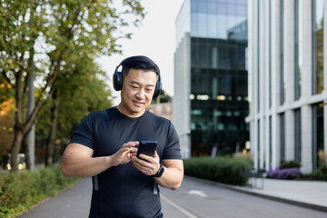 A young Asian man in sportswear is standing on a city street wearing headphones and using a mobile...