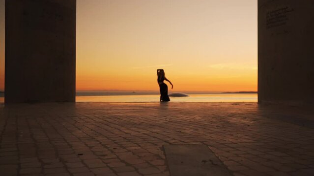 Woman dancing at sunset near water. Silhouette of lady performing graceful dance in rays of setting sun. Female dancer with long wavy hair moving body rhythmically in evening near river
