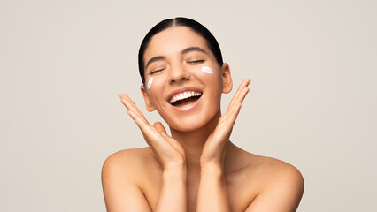 Beauty shot of gorgeous multiracial woman with amazing smile applying moisturizer on her face. Seasonal face skin protection