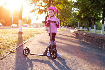 a beautiful girl in a pink helmet and protective equipment stands with a scooter on an alley in the city against the backdrop of a sunny sunset in summer.