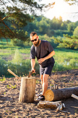 A man with a beard, a lumberjack with an ax in his hands, cuts wood in nature in the summer against the backdrop of a forest and a river bank. Camping. Survival, travel