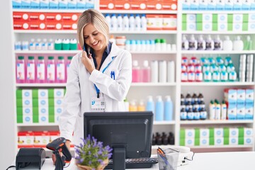 Young blonde woman pharmacist talking on smartphone using computer at pharmacy