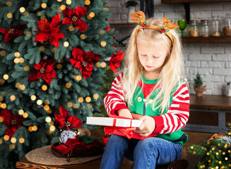 Child opening gift box with red ribbon in Christmas decorated kitchen. Kid in reindeer antlers with holly berries headband dressed up as elf. Xmas tree decorated with ornaments, tinsel and garland
