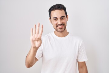 Handsome hispanic man standing over white background showing and pointing up with fingers number four while smiling confident and happy.