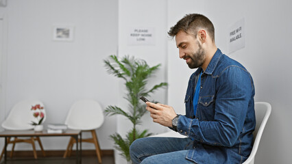 Smiling young hispanic man relaxes by typing message on smartphone screen, sitting in the chair of...
