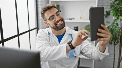 Young hispanic man doctor making selfe picture by touchpad smiling at the clinic