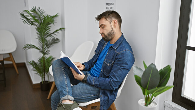 Relaxed young hispanic male settling into a waiting room chair, engrossed in a book - a handsome portrait of rest, focus, and ongoing pursuit of knowledge. indoor corridor forms a serene background.