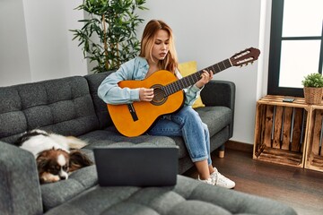 Young caucasian woman having online classical guitar class sitting on sofa with dog at home