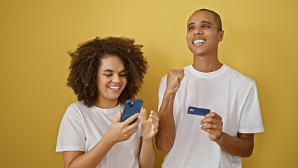 Smiling beautiful couple in love, celebrating by shopping online, confidently using credit card on smartphone over a yellow, isolated background