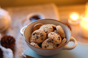 Bowl of cookies, cup of tea or coffee, plate of blueberries, dry oranges, wool yarn, pine cones, books, reading glasses, tablet, pen, soft blanket and lit candles on the table. Winter hygge at home.