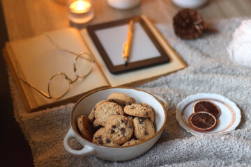 Bowl of cookies, cup of tea or coffee, plate of blueberries, dry oranges, wool yarn, pine cones, books, reading glasses, tablet, pen, soft blanket and lit candles on the table. Winter hygge at home.
