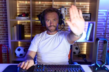 Middle age man with beard playing video games wearing headphones doing stop sing with palm of the...