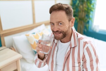 Middle age man drinking glass of water sitting on bed at bedroom