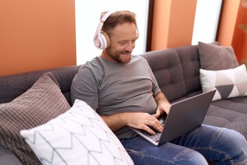 Middle age man using laptop and headphones sitting on sofa at home