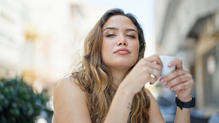 Fototapeta premium Young beautiful hispanic woman sitting on table holding cup coffee at coffee shop terrace