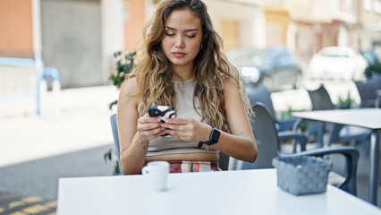 Young beautiful hispanic woman using smartphone sitting on table at coffee shop terrace