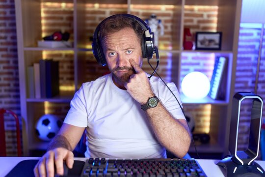 Middle age man with beard playing video games wearing headphones pointing to the eye watching you gesture, suspicious expression