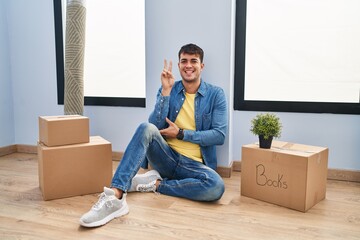 Young hispanic man sitting on the floor at new home smiling with happy face winking at the camera...