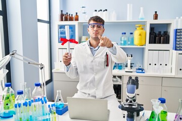 Young hispanic man working at scientist laboratory holding diploma pointing with finger to the...