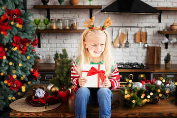 Child holding Christmas present in festive decorated kitchen. Kid in reindeer antlers with holly...