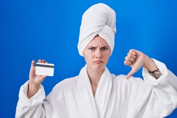 Blonde caucasian woman wearing bathrobe holding credit card with angry face, negative sign showing dislike with thumbs down, rejection concept