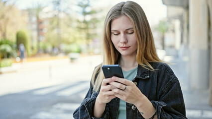Young blonde woman using smartphone with serious face at street