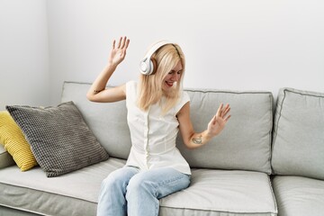 Obraz premium Young blonde woman listening to music sitting on sofa dancing at home