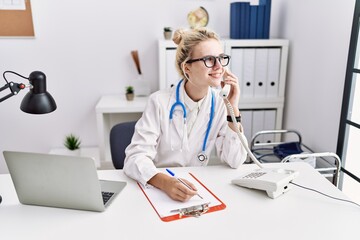 Young blonde woman doctor talking on telephone writing on document at clinic