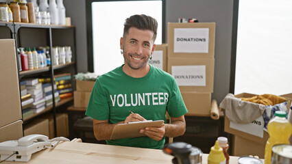 Attractive young hispanic man volunteering with a smile, writing on clipboard at charity center amidst donations