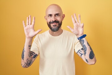 Hispanic man with tattoos standing over yellow background showing and pointing up with fingers number nine while smiling confident and happy.