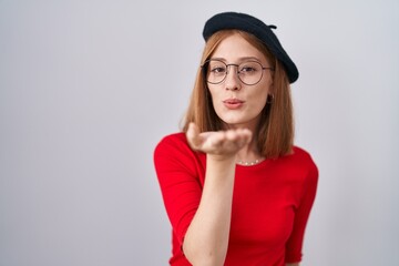 Young redhead woman standing wearing glasses and beret looking at the camera blowing a kiss with hand on air being lovely and sexy. love expression.