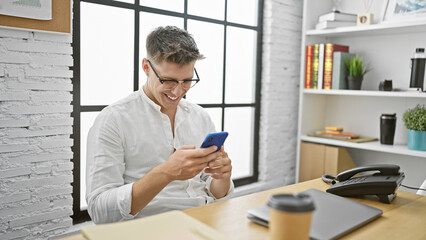 Hardworking young caucasian man in business, beaming while typing a message via smartphone at the office desk.