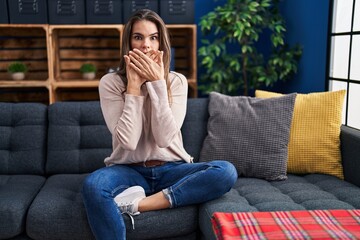 Beautiful woman sitting on the sofa at home shocked covering mouth with hands for mistake. secret...