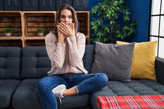 Beautiful woman sitting on the sofa at home shocked covering mouth with hands for mistake. secret concept.